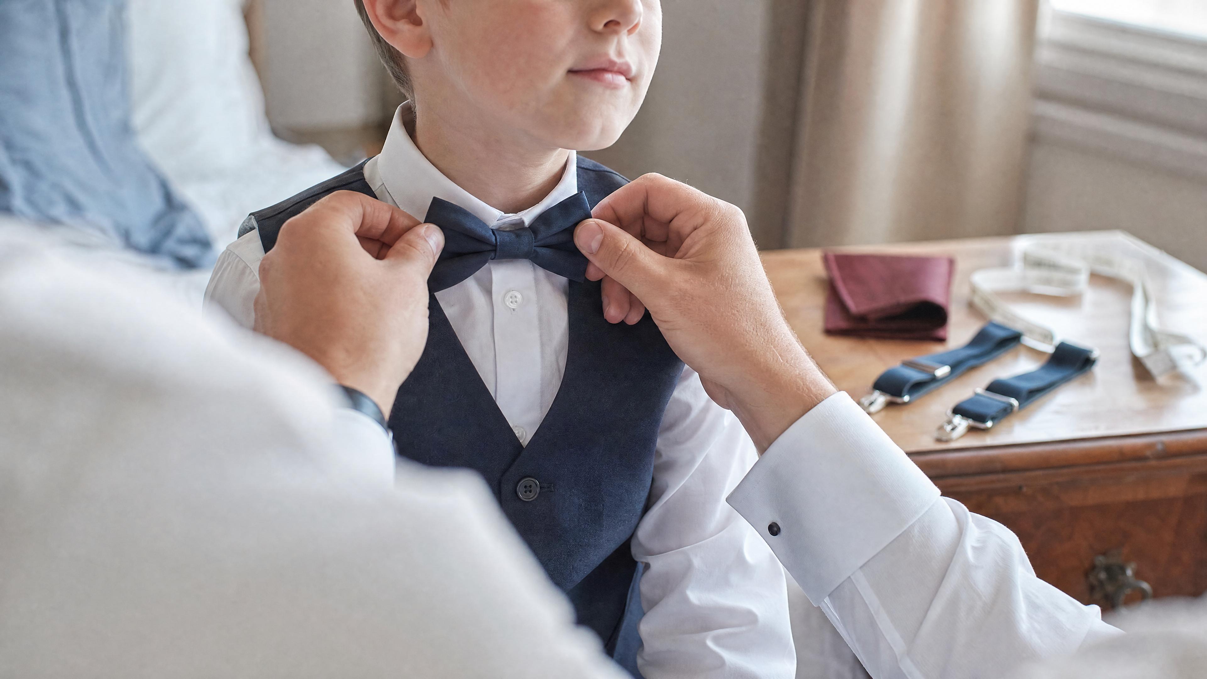 Un adulte ajuste un nœud papillon sur un enfant en chemise blanche, avec un mètre ruban visible sur une commode, dans une chambre lumineuse avant une cérémonie Bild